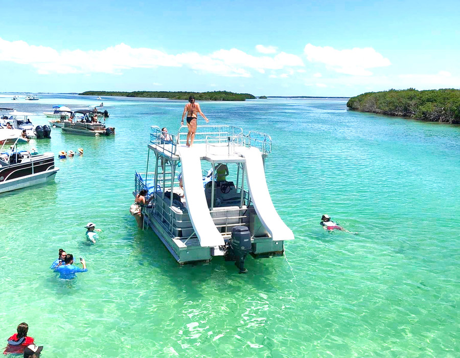 A double-slide pontoon boat anchored in turquoise shallows with swimmers around it